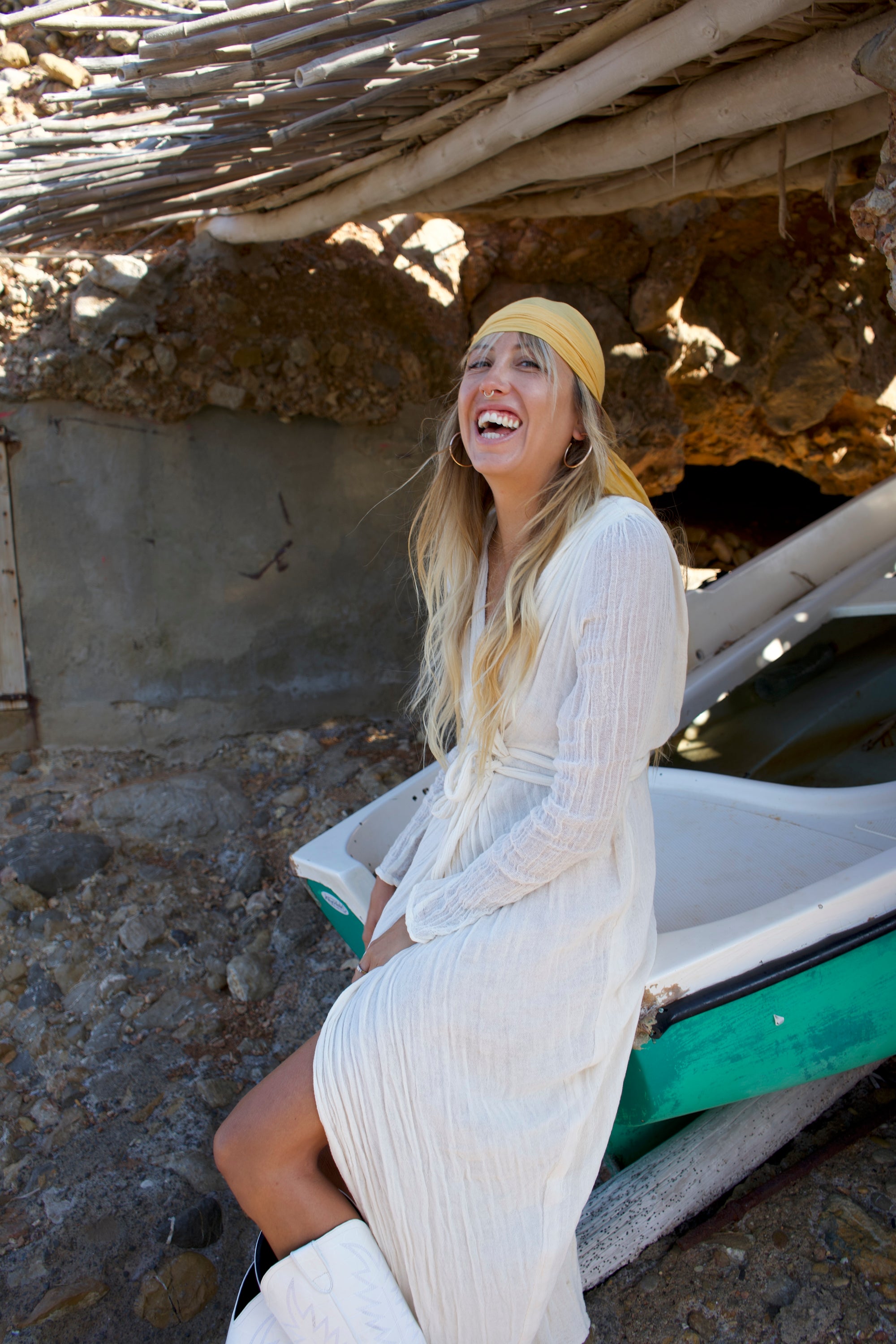 Woman in a white dress and yellow headscarf sitting in a small boat near a stone wall.